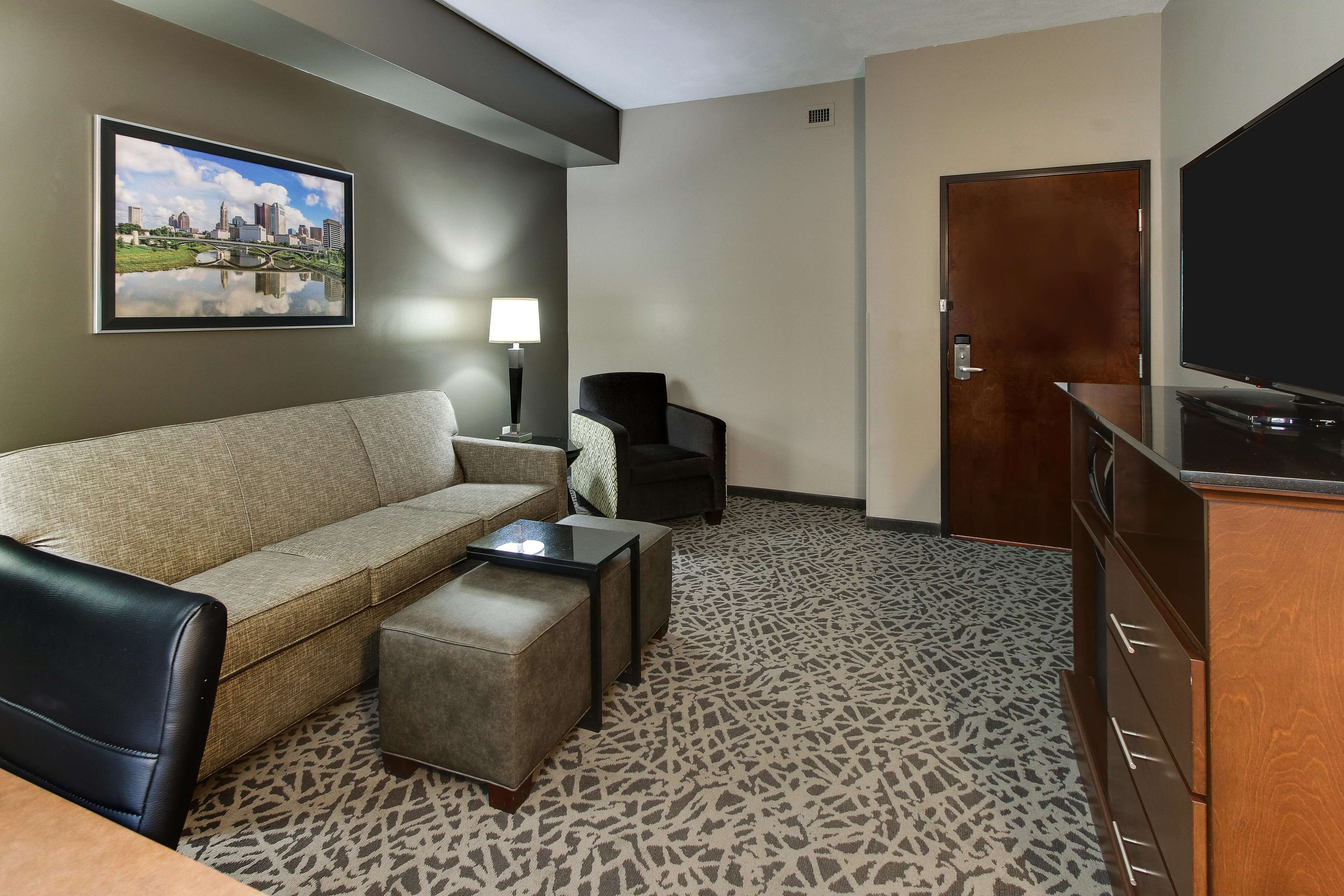 A hotel room with a sofa, a chair, a small table, a TV, and a door.