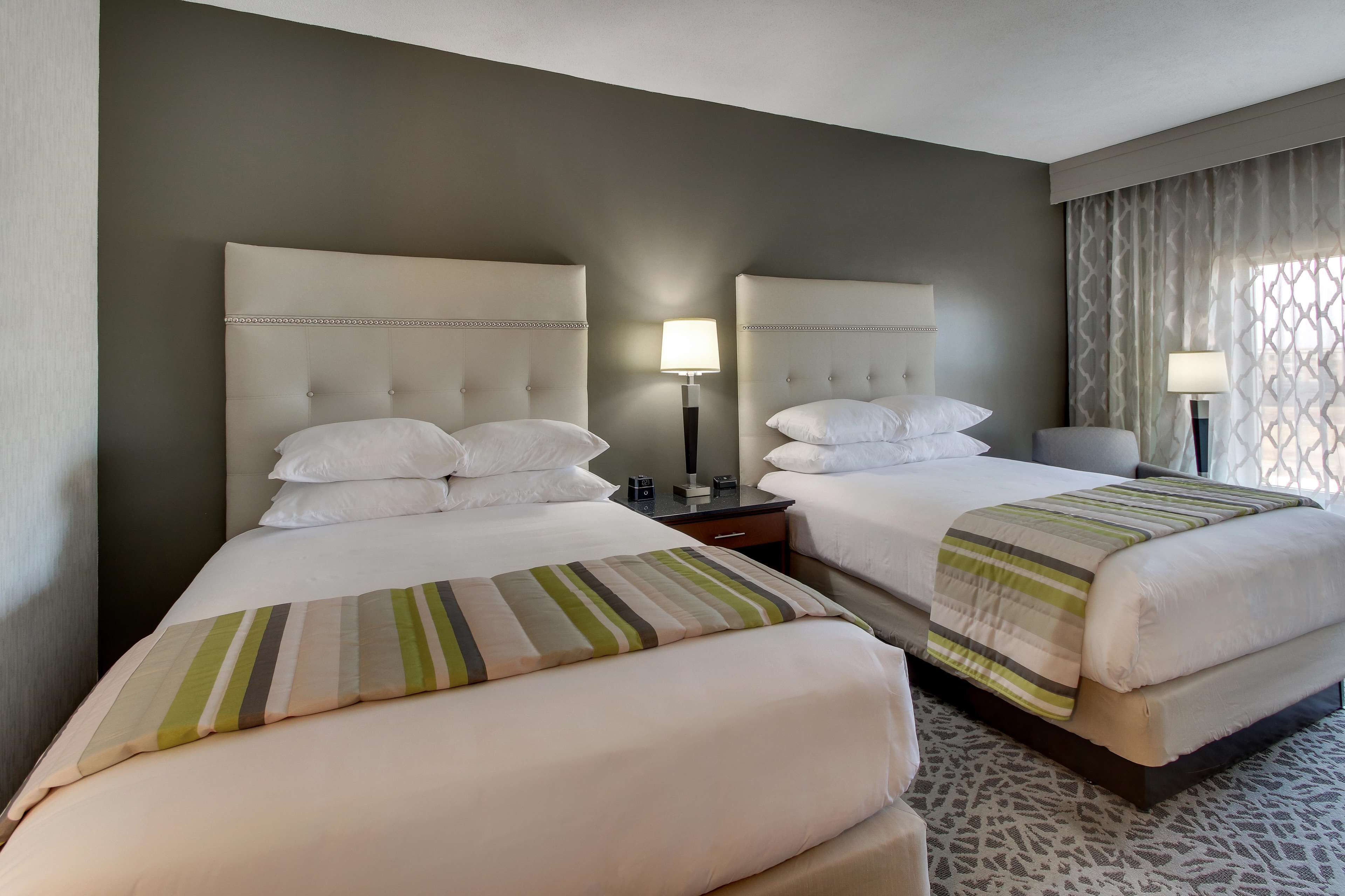 A hotel room with two beds, each with a headboard, a bedside table, a lamp, and a patterned rug.