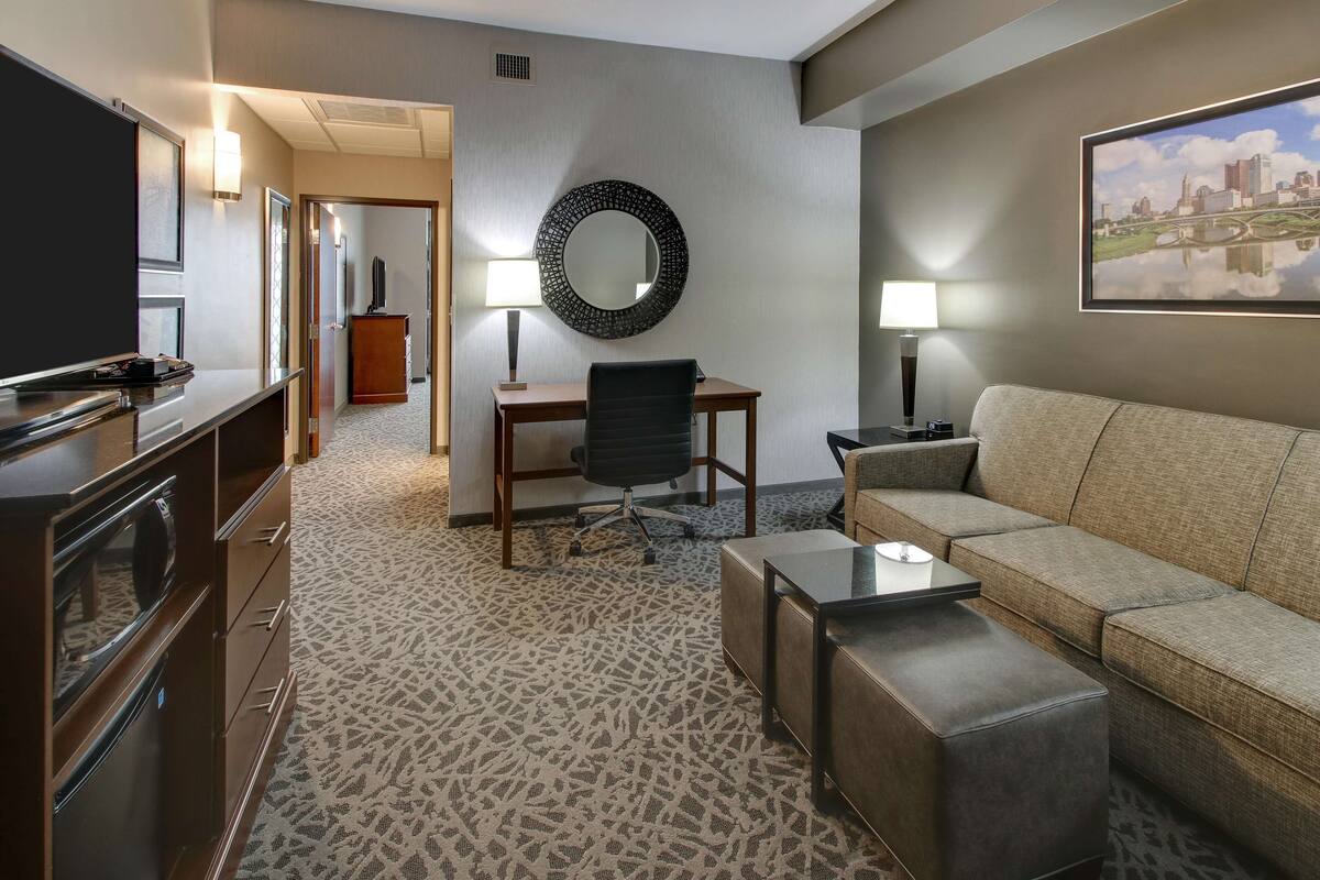 A hotel room with a sofa, a TV, a desk with a chair, a mirror, and a patterned carpet.