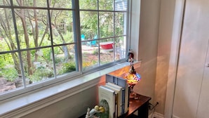 A room with a window overlooking a garden, a small table with books and a figurine, and a decorative lamp. - Garden Cottage, Far From The Maddening Crowd but so close to the night life! (Dallas)