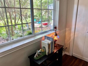 A room with a window overlooking a garden, a small table with books and a figurine, and a decorative lamp.