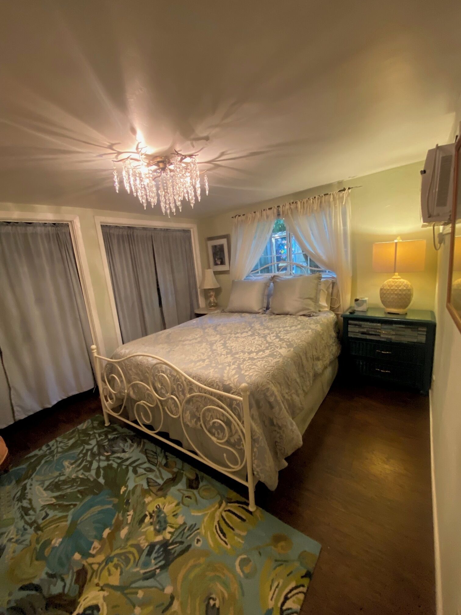 A bedroom with a bed, curtains, a chandelier, a nightstand, and a lamp.