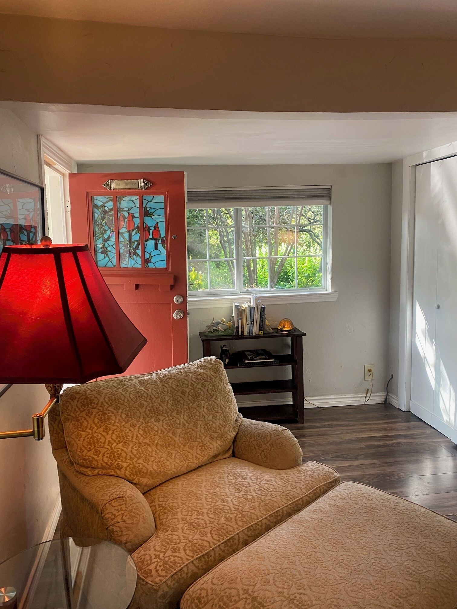 A room with a patterned armchair, a red lamp, a bookshelf with books and decorative items, and a window with a view of greenery.