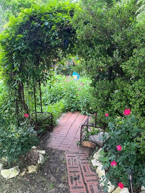A garden pathway with a brick pattern, surrounded by greenery and flowering plants.
