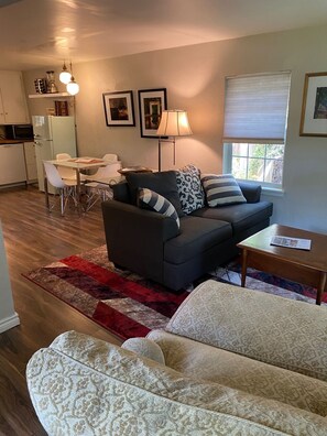 A living room with a dark sofa, a patterned rug, a white couch, and a dining area with a table and chairs.