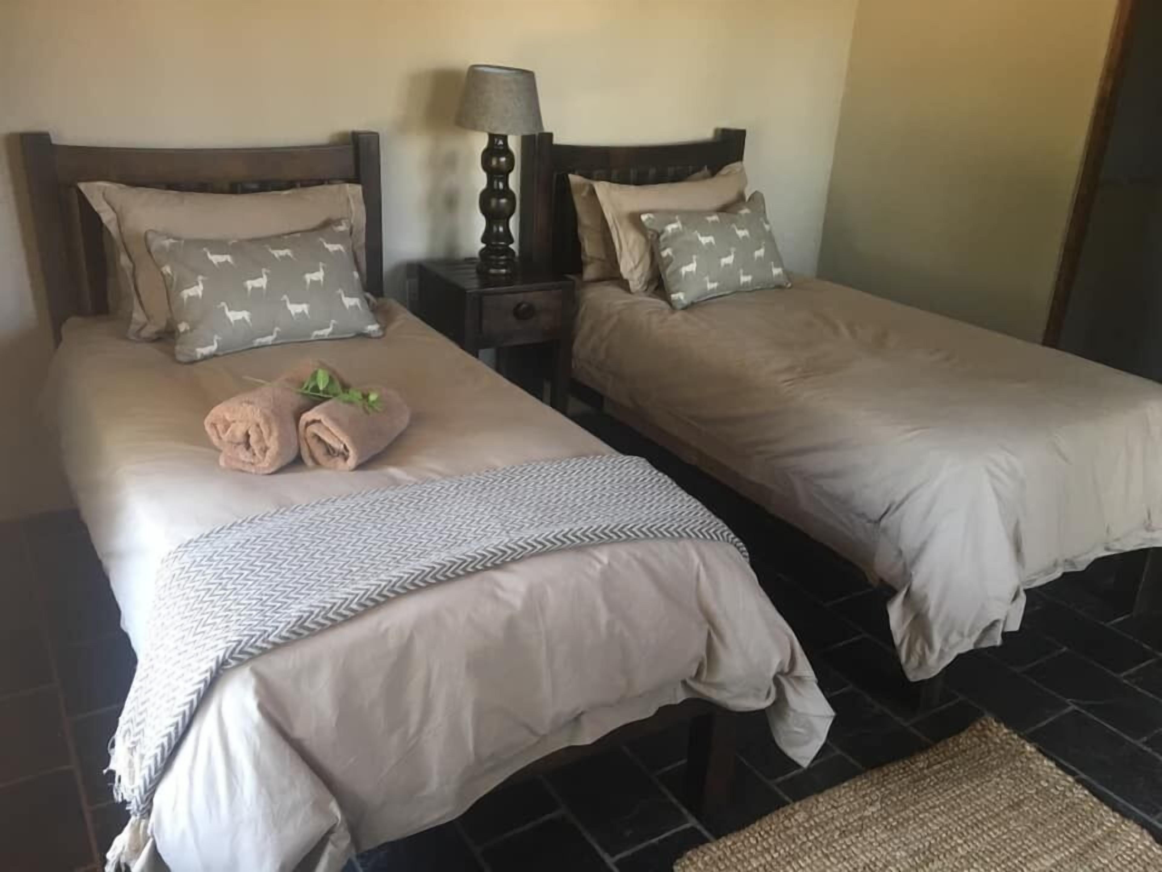 Habitación de hotel con dos camas individuales, cada una con almohada y manta. Se observa un mesita de noche con lámpara entre las camas.