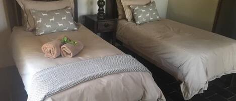 Habitación de hotel con dos camas individuales, cada una con almohada y manta. Se observa un mesita de noche con lámpara entre las camas.