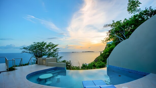 Una piscina a sfioro con vista sul mare, circondata da lettini prendisole e un tavolino.