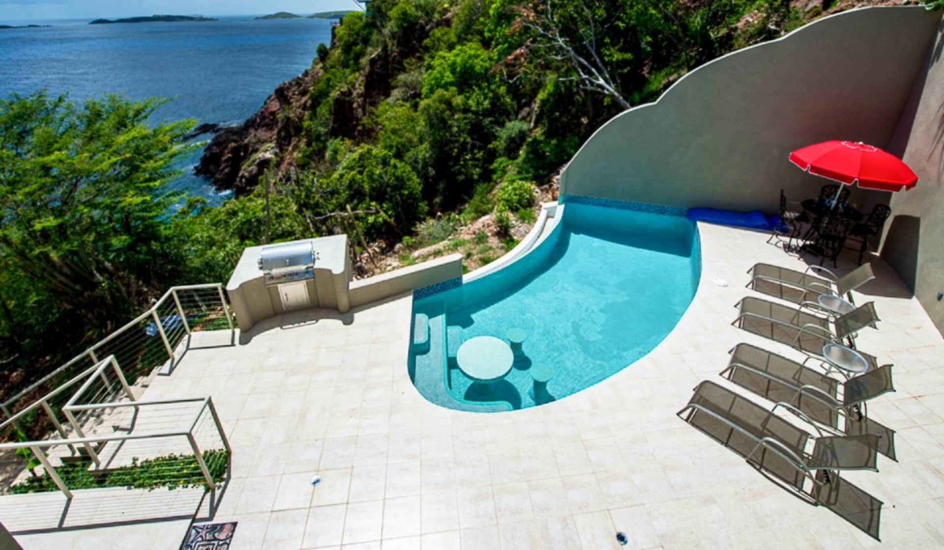 A swimming pool with a curved edge, surrounded by lounge chairs and a red umbrella.