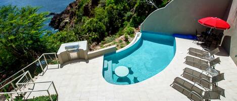 A swimming pool with a curved edge, surrounded by lounge chairs and a red umbrella.