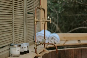 Une salle de bain avec une baignoire en bois, une fenêtre avec des stores et une rangée de produits de toilette, notamment du shampoing et de l’après-shampoing.