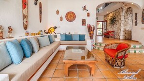Ein großzügiger Wohnbereich mit einer Couch, einem Couchtisch und einem Korbstuhl. Der Raum verfügt über eine Steinwand und eine Treppe, die zu einer oberen Ebene führt.
