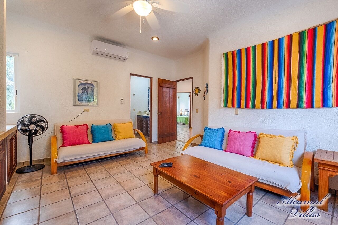 Un salon avec un canapé, une table basse en bois et un ventilateur. On y trouve une décoration murale colorée à rayures et un tableau encadré.