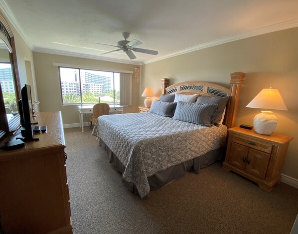 A bedroom with a bed, a desk, a chair, a nightstand, a lamp, a ceiling fan, and a window with a view of buildings. - SIESTA DUNES 1-406 LUXURY CONDO WITH A GREAT VIEW! (Sarasota)
