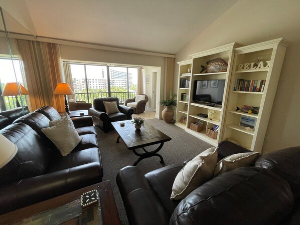 A living room with leather sofas, a coffee table, a bookshelf, and a television. - SIESTA DUNES 1-406 LUXURY CONDO WITH A GREAT VIEW! (Sarasota)
