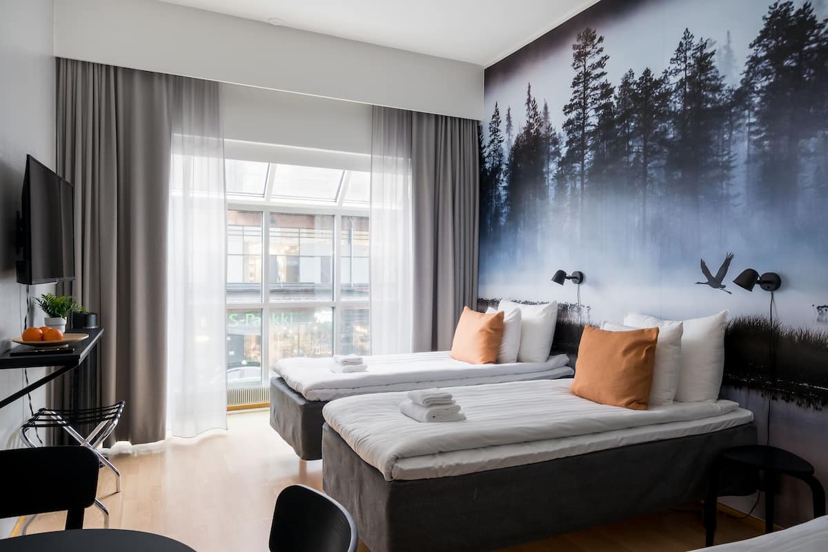A modern hotel room with two beds, a desk with a TV, and a large window with curtains.