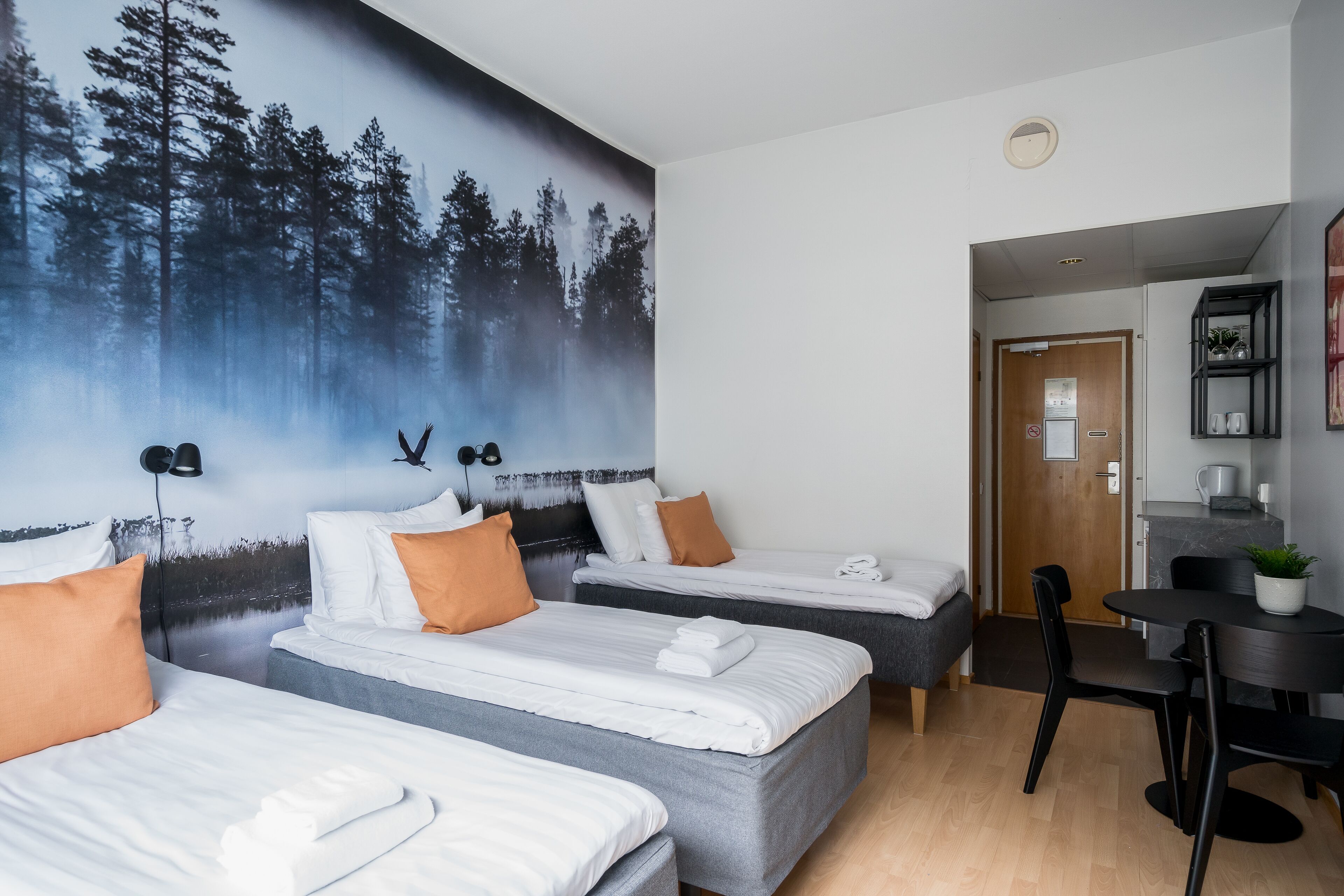 Habitación de hotel con tres camas, una mesa de comedor y un mural de un paisaje forestal.
