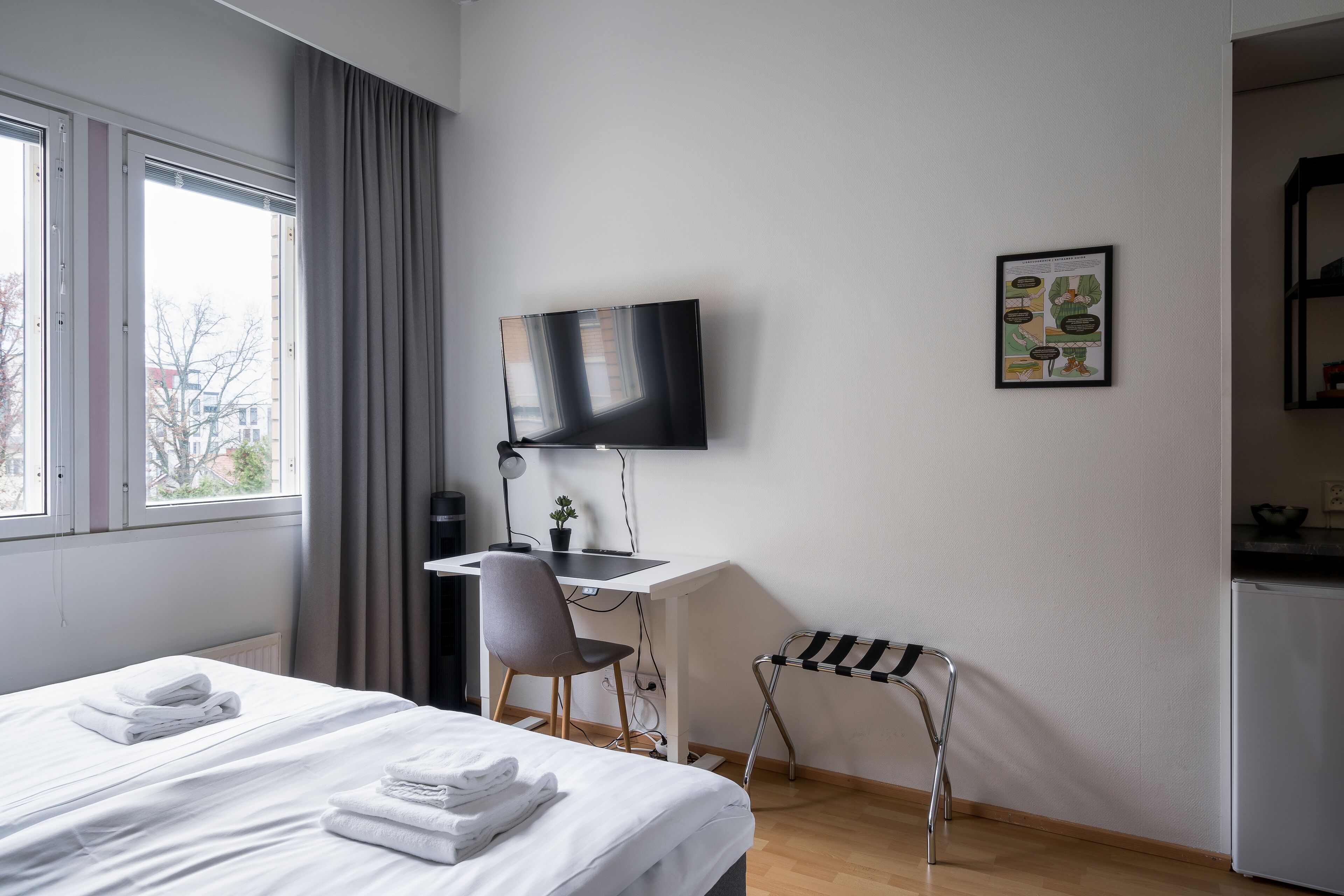 A hotel room with a bed, a desk, a chair, a TV, and a small table with a plant.