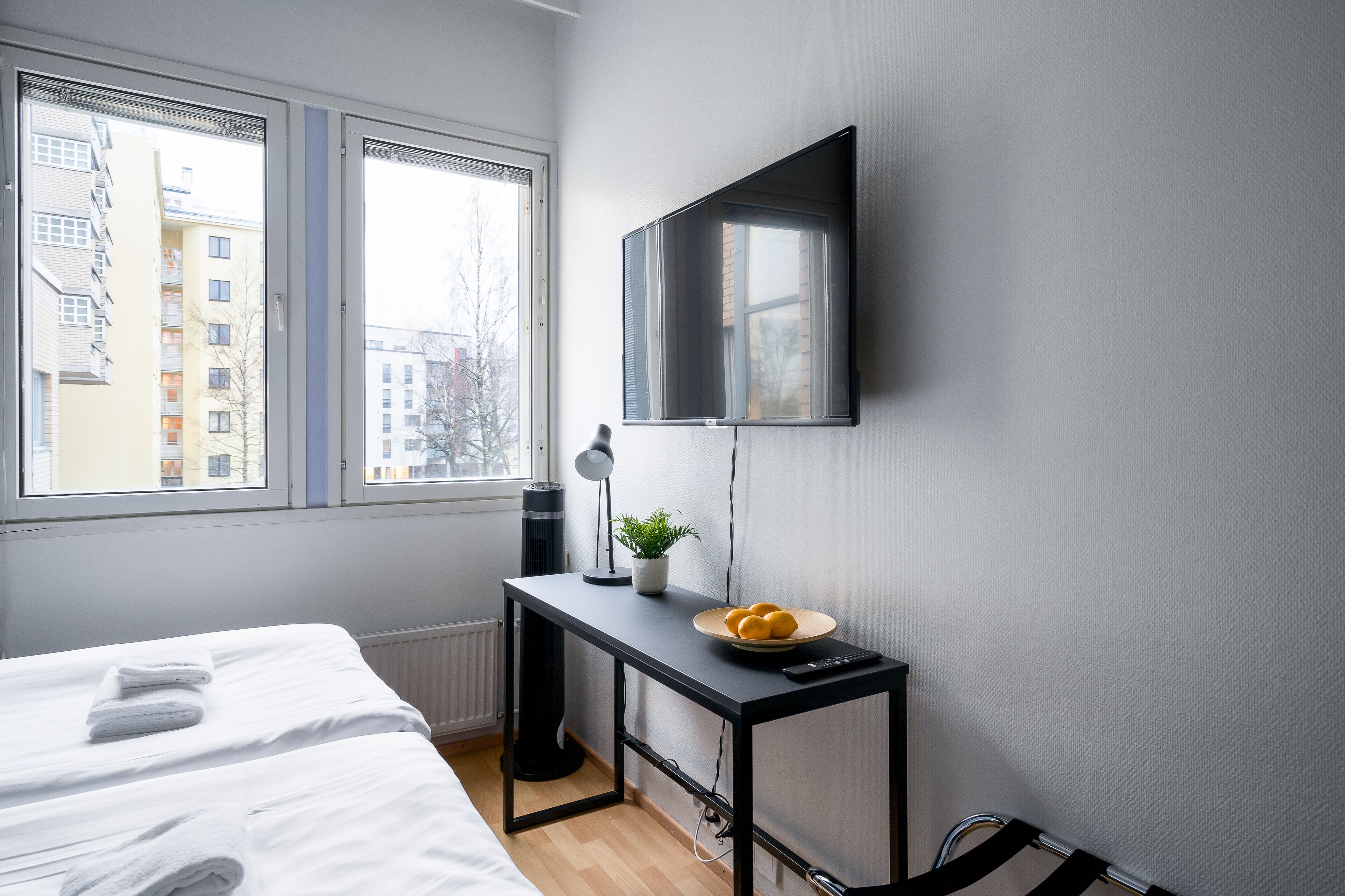 A bedroom with a bed, a desk with a plant, a bowl of fruit, and a TV on the wall.