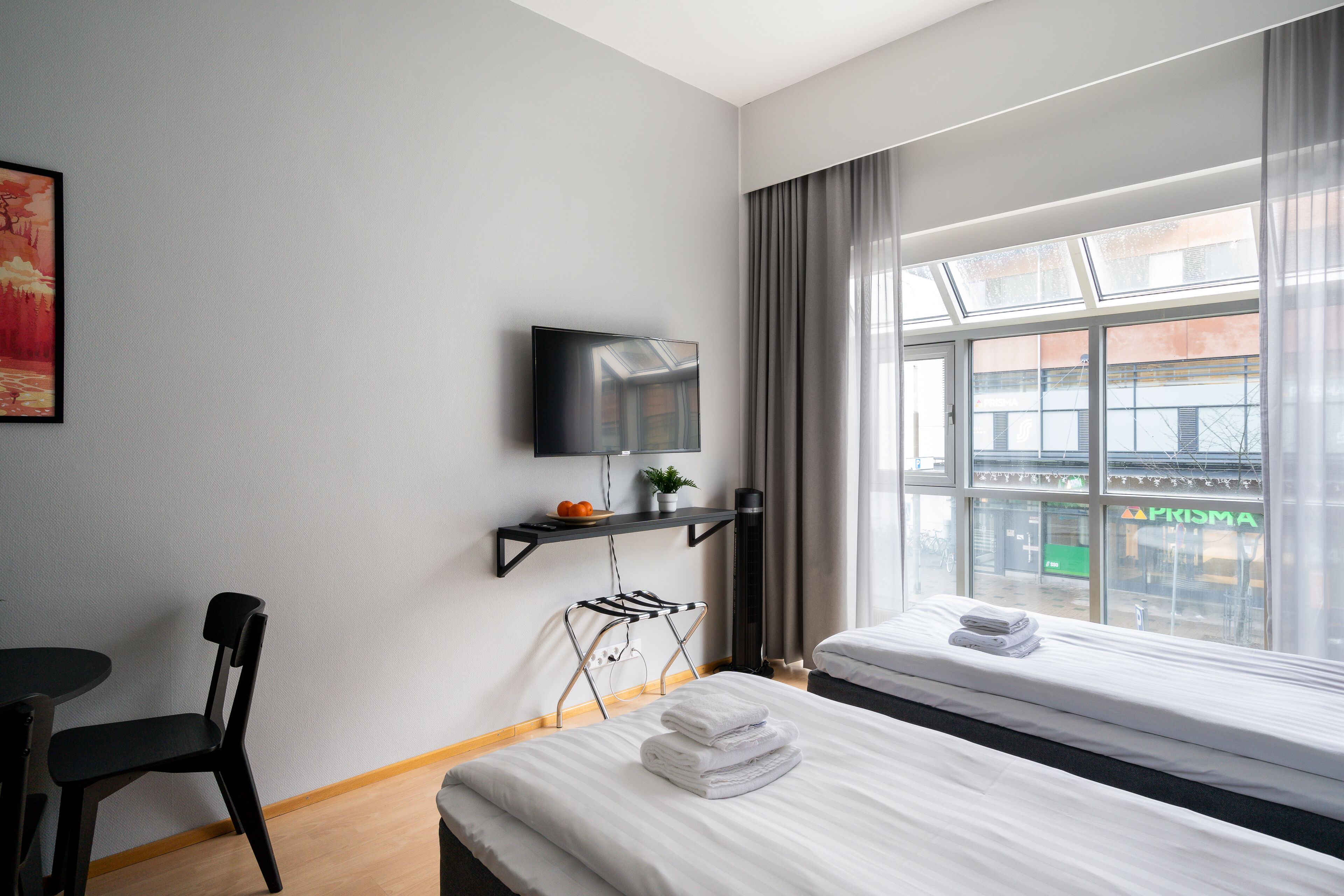 Et hotelværelse med seng, stol, fjernsyn ophængt på væggen, et vindue med gardiner og en lille hylde med en plante og en skål med frugt.