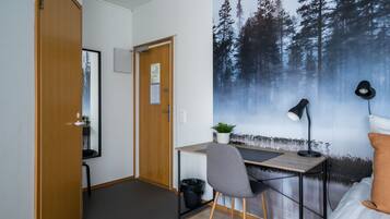 Una habitación con una cama, un escritorio con una silla, una computadora, una lámpara y un gran mural de un bosque en la pared.