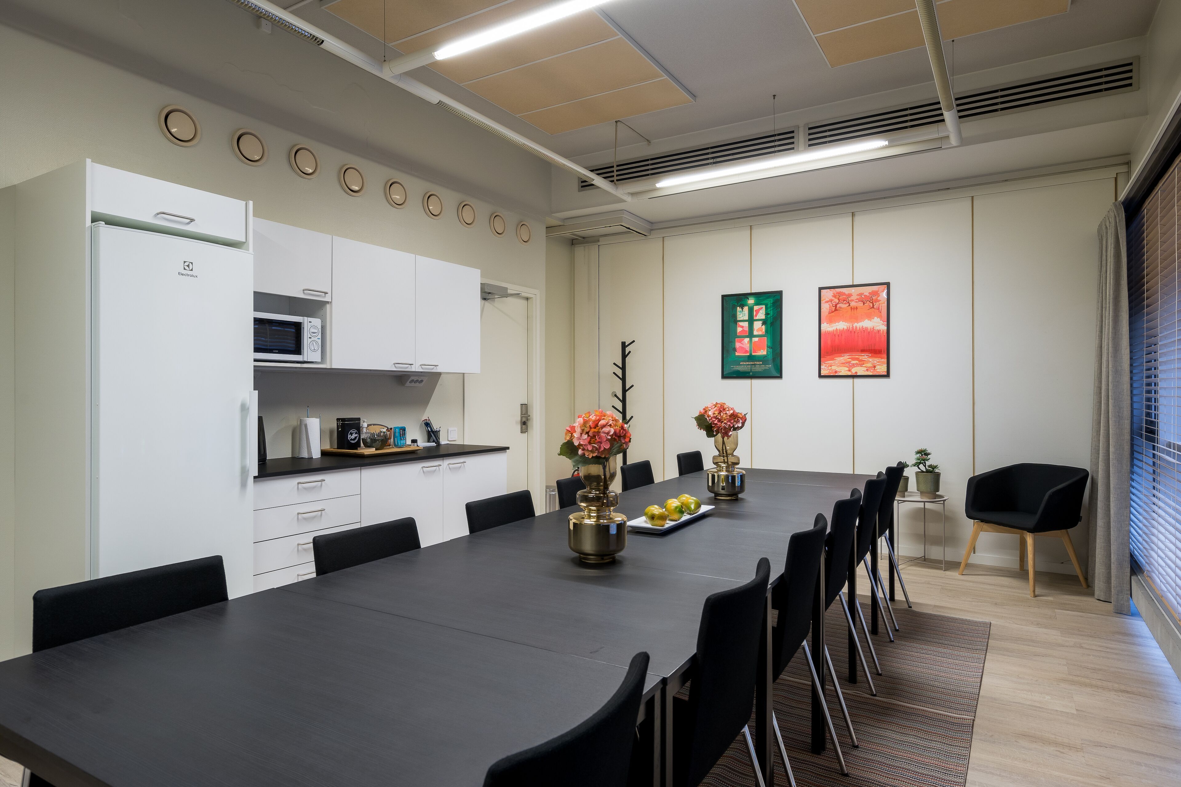 A modern conference room with a long table, black chairs, a white refrigerator, a microwave, and a small kitchen area.