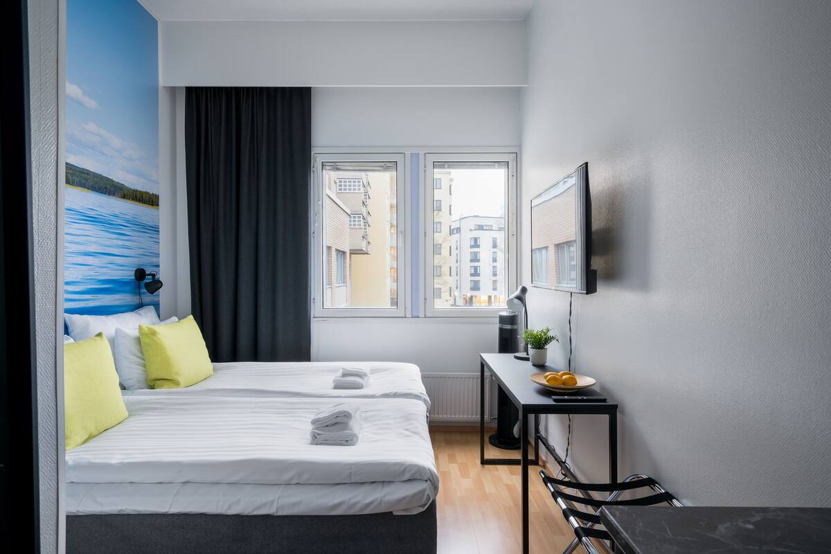 A hotel room with two beds, a desk, a chair, a window with a view of buildings, and a wall-mounted TV.