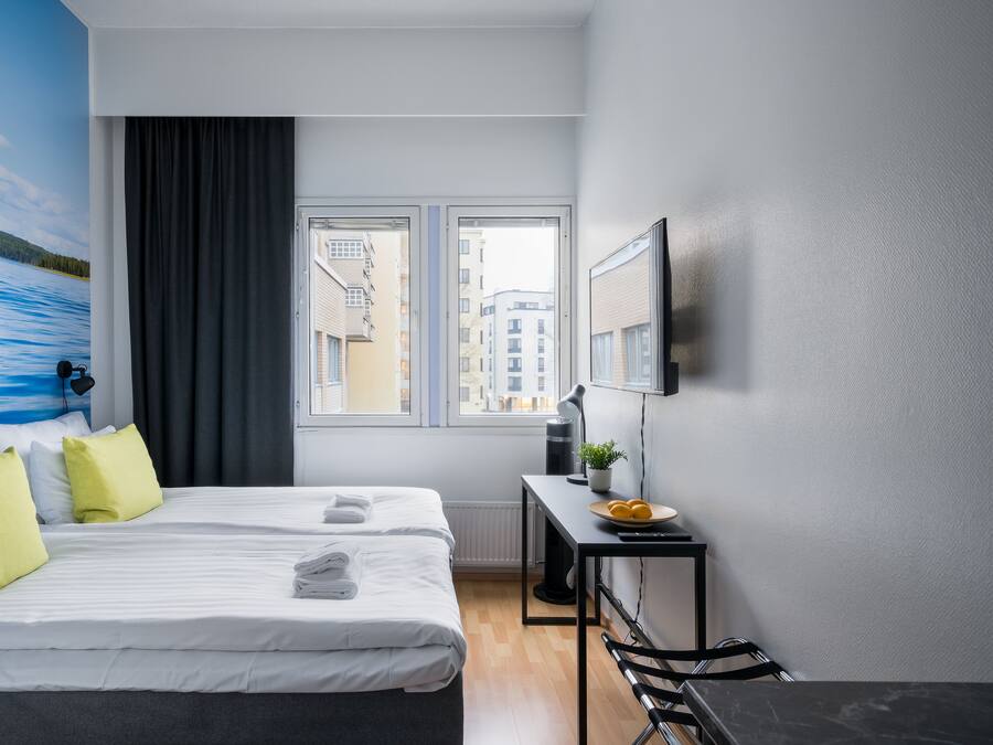 Quarto de hotel com duas camas, uma escrivaninha, uma cadeira, janela com vista para prédios e TV na parede.