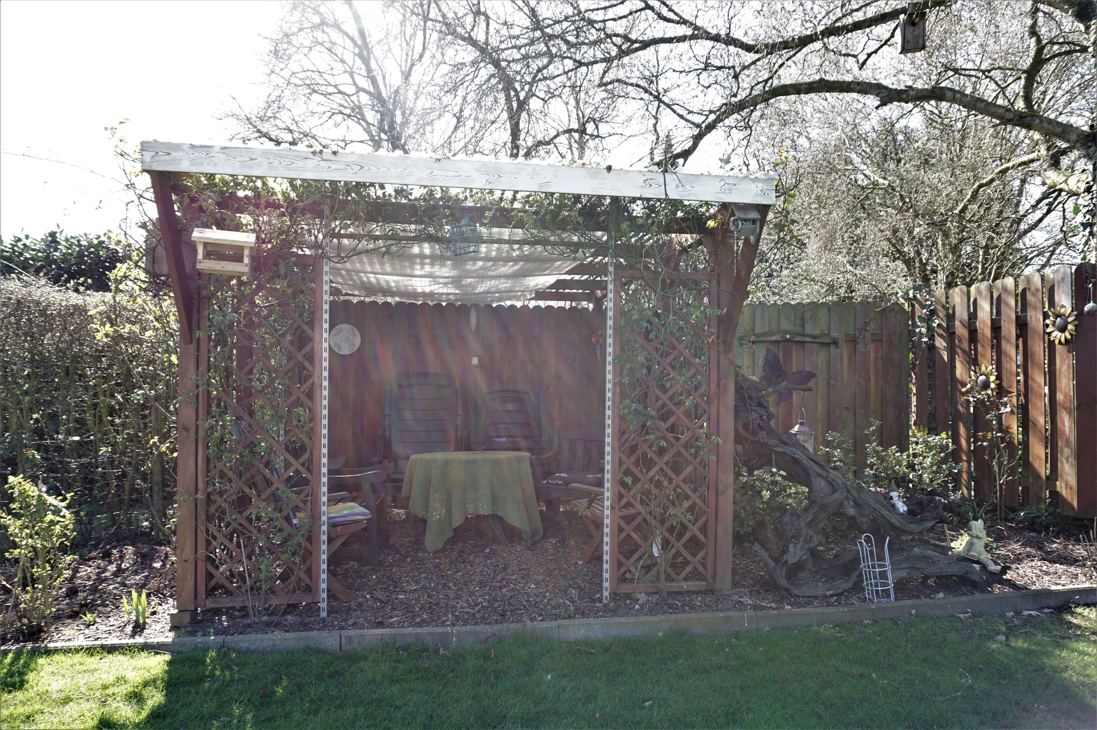 A wooden pergola with a table and chairs underneath, surrounded by a garden with a wooden fence and trees.