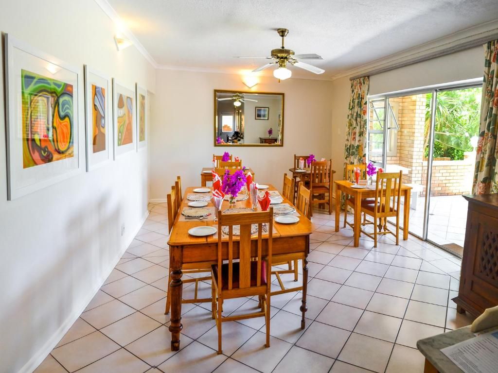 Un coin repas avec une grande table dressée pour quatre personnes, entourée de chaises. Un ventilateur de plafond est installé au-dessus, et une grande fenêtre avec des rideaux se trouve sur le côté droit.