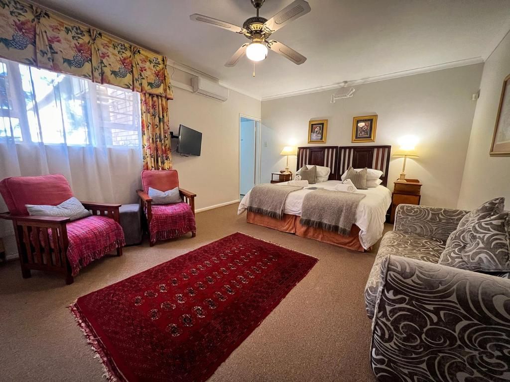 Habitación de hotel con cama, dos sillones, un sofá, alfombra roja, ventilador de techo y televisión en la pared.