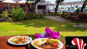 An outdoor dining area with tables, umbrellas, and a view of a building with a corrugated metal roof. - Sundune Guest House & Self Catering (Colchester)