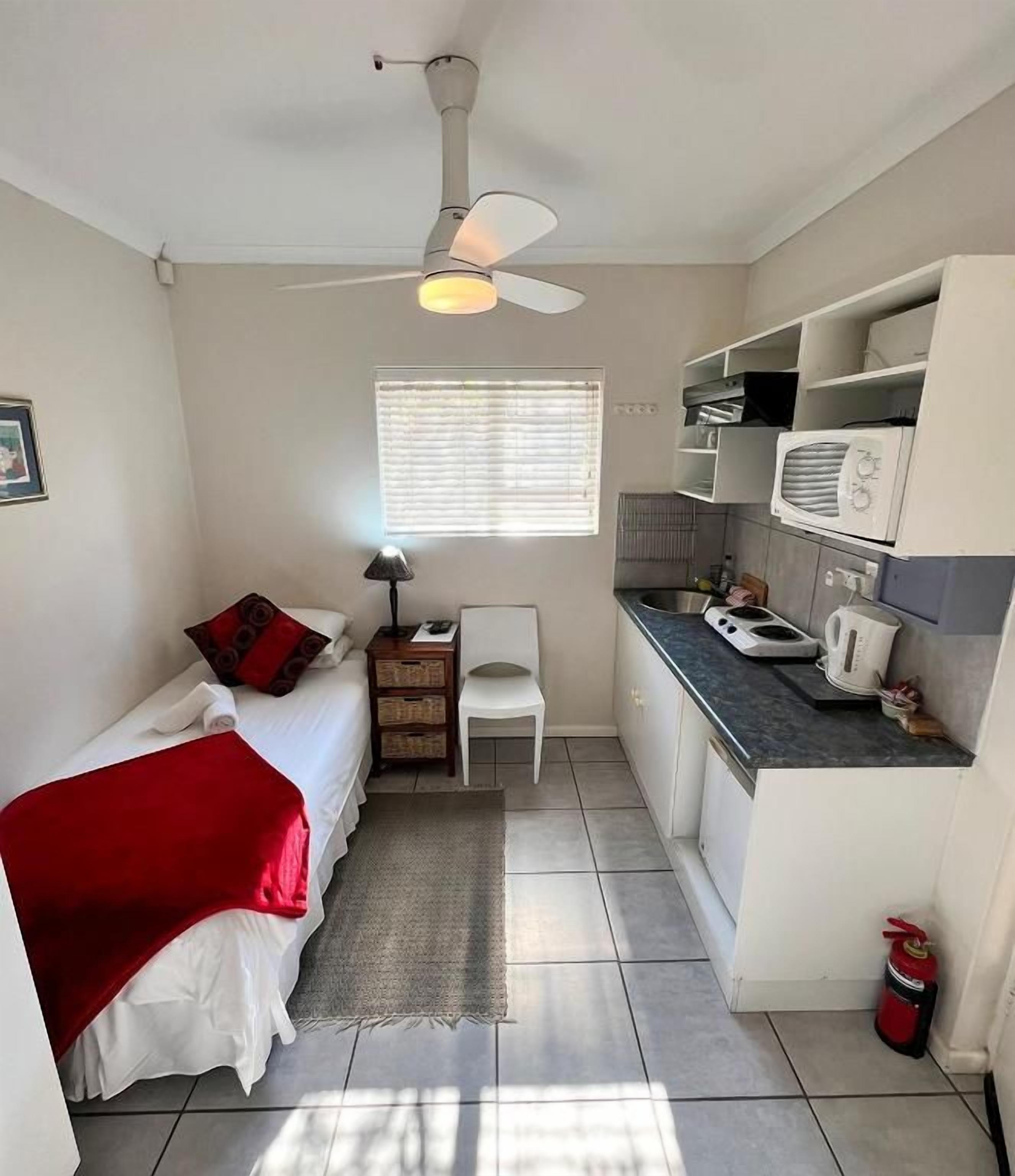 A compact room with a bed, a desk, a chair, a kitchenette, and a ceiling fan.