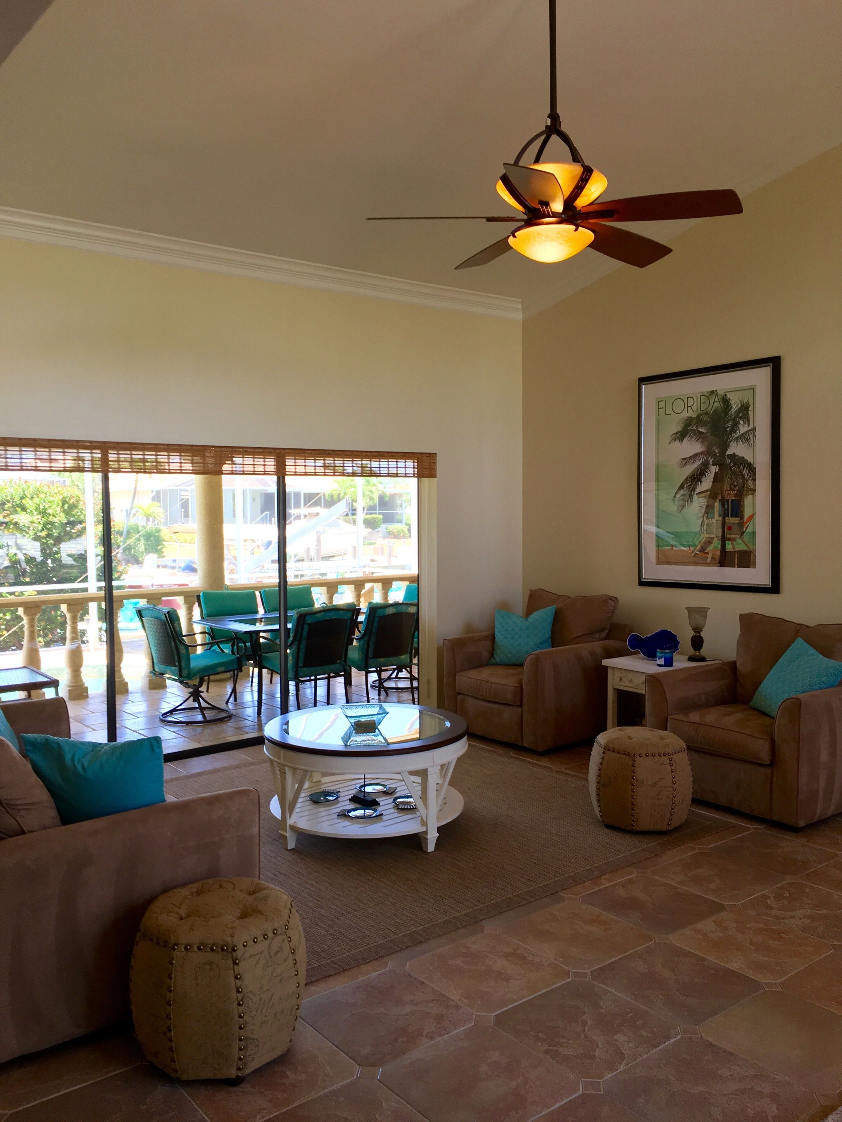 A spacious living area with a ceiling fan, a glass coffee table, and a view of an outdoor patio with green chairs and a table.