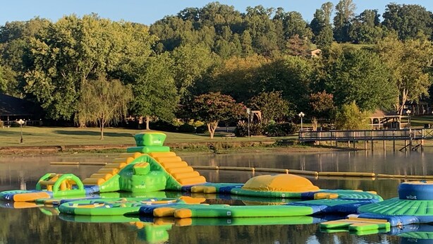 Ein Wasserspielbereich mit aufblasbaren Elementen und einer Brücke über einen stillen See, umgeben von Bäumen und einem Pavillon.