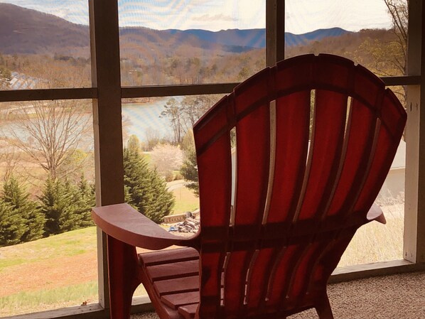 A red Adirondack chair facing a scenic view through large windows, including a lake and mountains.