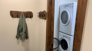 Een wasserette met een wasmachine en droger, een houten kapstok met een jas, en een zwarte mat op de vloer.