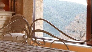 Une pièce avec un lit, une chaise en osier et une vue sur une colline boisée, visible par la fenêtre.