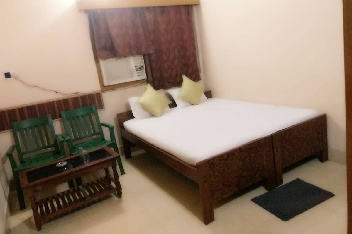 A room with a bed, two green chairs, a small table, a window with curtains, and a wall-mounted air conditioner.