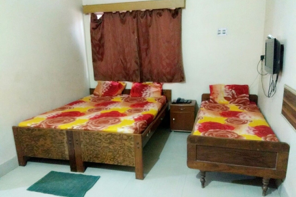 A room with two wooden beds, a small bedside table, a TV mounted on the wall, and a window with curtains.