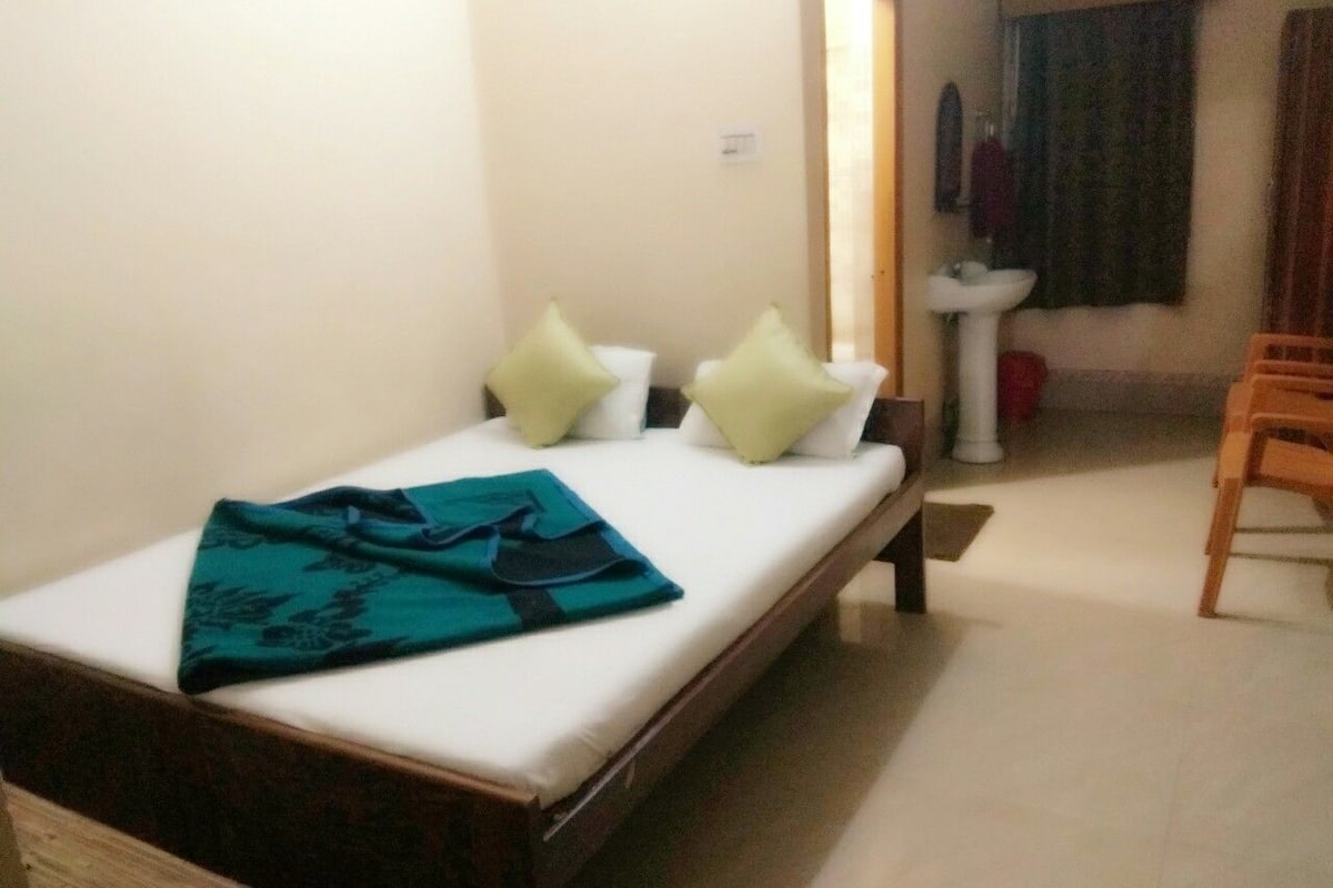 A single bed with a teal blanket, two pillows, a bedside table, and a wooden chair in a room with a white sink and a mirror on the wall.