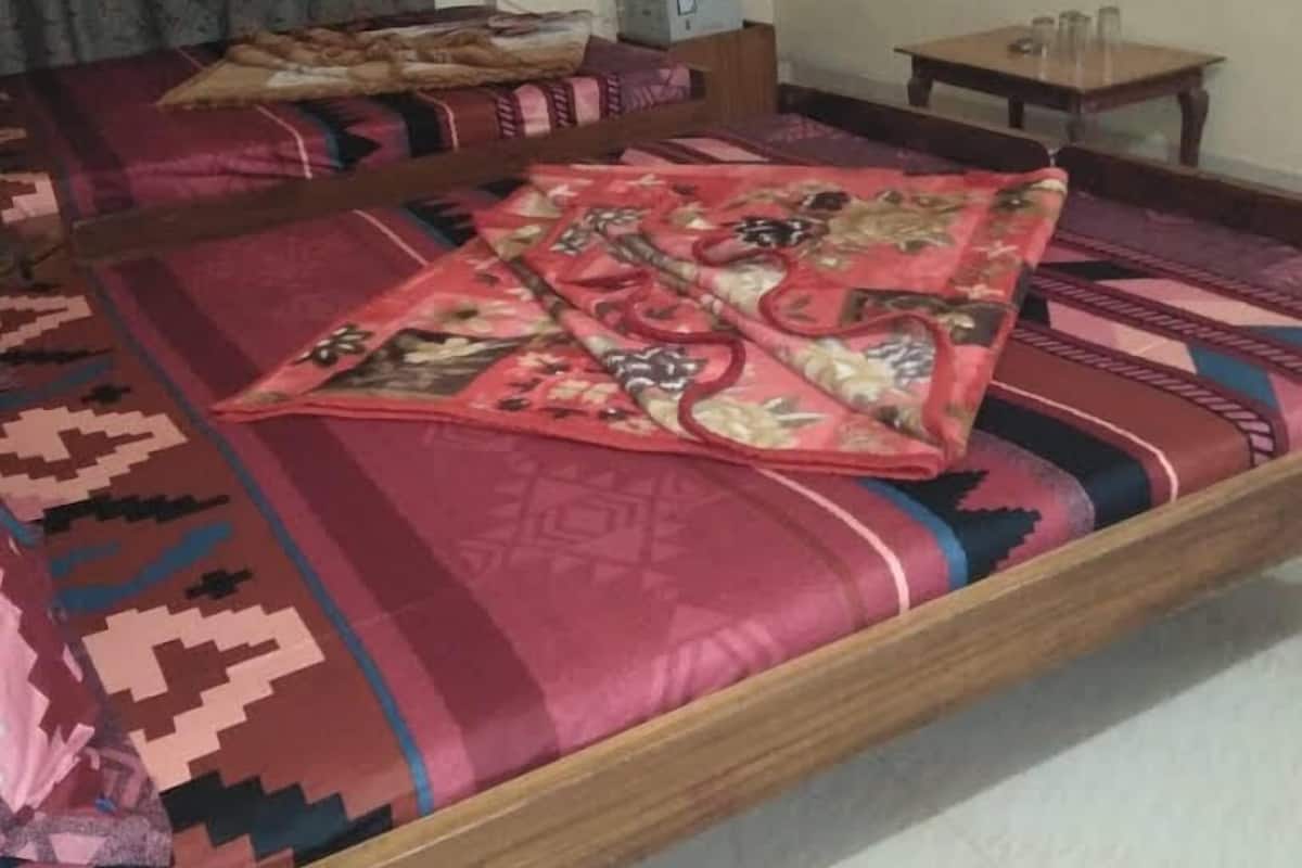 A bedroom with a bed covered in patterned bedding, a wooden headboard, a bedside table with a lamp, and a wall-mounted TV.