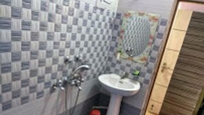 A bathroom with a checkered tile wall, a sink, a mirror, and a shower. - Hotel Anjali (Deoghar)