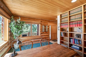 Ein Holzinnenraum mit Esstisch, Stühlen, einem Bücherregal und einem Fenster mit Blick auf Bäume.