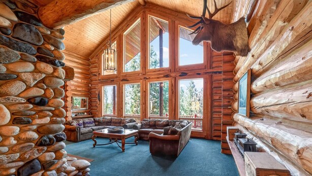 A log cabin interior with a stone wall, large windows, a fireplace, and a mounted elk head. - Luxury Log Cabin Near Cascade Lake, 80 Acres, RVs (Cascade)