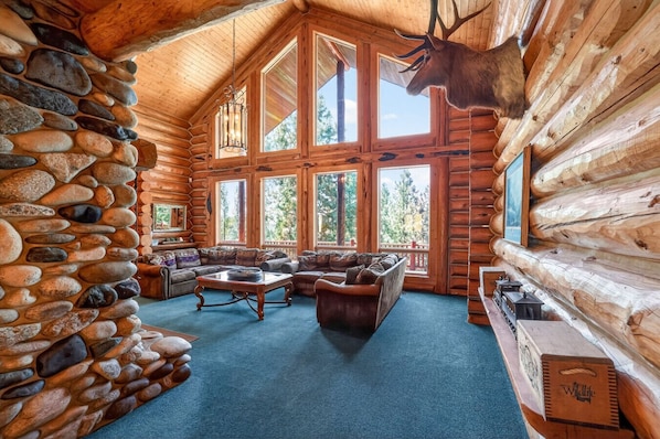 A log cabin interior with a stone wall, large windows, a fireplace, and a mounted elk head.