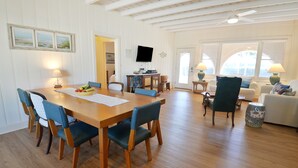 Un vaste espace de vie comprenant une table à manger en bois, des chaises bleues, un téléviseur et un coin salon confortable avec un canapé blanc et un pouf à motifs.