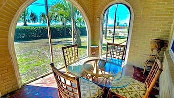 Relax in a room with a glass table and comfortable chairs, surrounded by arched windows offering a view of the lush lawn and palm trees.