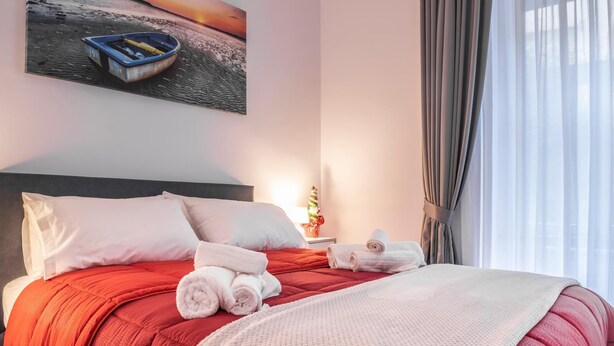 A neatly made bed with red bedding, white pillows, and rolled towels. A painting of a boat on water is on the wall, and a small Christmas tree is on the nightstand. - Casina Rossa Apartment Procida (Napoli)