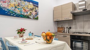 A modern kitchen with a gas stove, oven, and wooden cabinets. A dining table with chairs and a vase of fruit is set up in front of a colorful cityscape painting. - Casina Rossa Apartment Procida (Napoli)